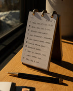 A Leather Discbound Analog + 3 Refills (Black) sits atop a desk, its full-grain leather cover open to reveal a handwritten to-do list with scheduled tasks. A pen etched with "UGMONK" rests in front of the pad, while warm sunlight pours through a nearby window, casting soft shadows across the workspace.