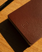 A Leather Discbound Analog (Brown) notebook crafted from full-grain leather rests on a wooden surface, its small embossed logo subtly visible. The top left corner reveals part of a black pen with gold text, suggesting it's used for jotting down quick Analog Task Cards. Sunlight casts a shadow across the scene.