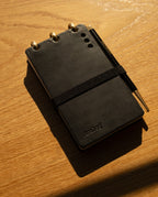 A Leather Discbound Analog + 3 Refills (Black) notebook, featuring full-grain black leather covers and an elastic band, sits on a wooden surface in warm light. A matching black pen rests on top, with Analog Task Cards visible inside.