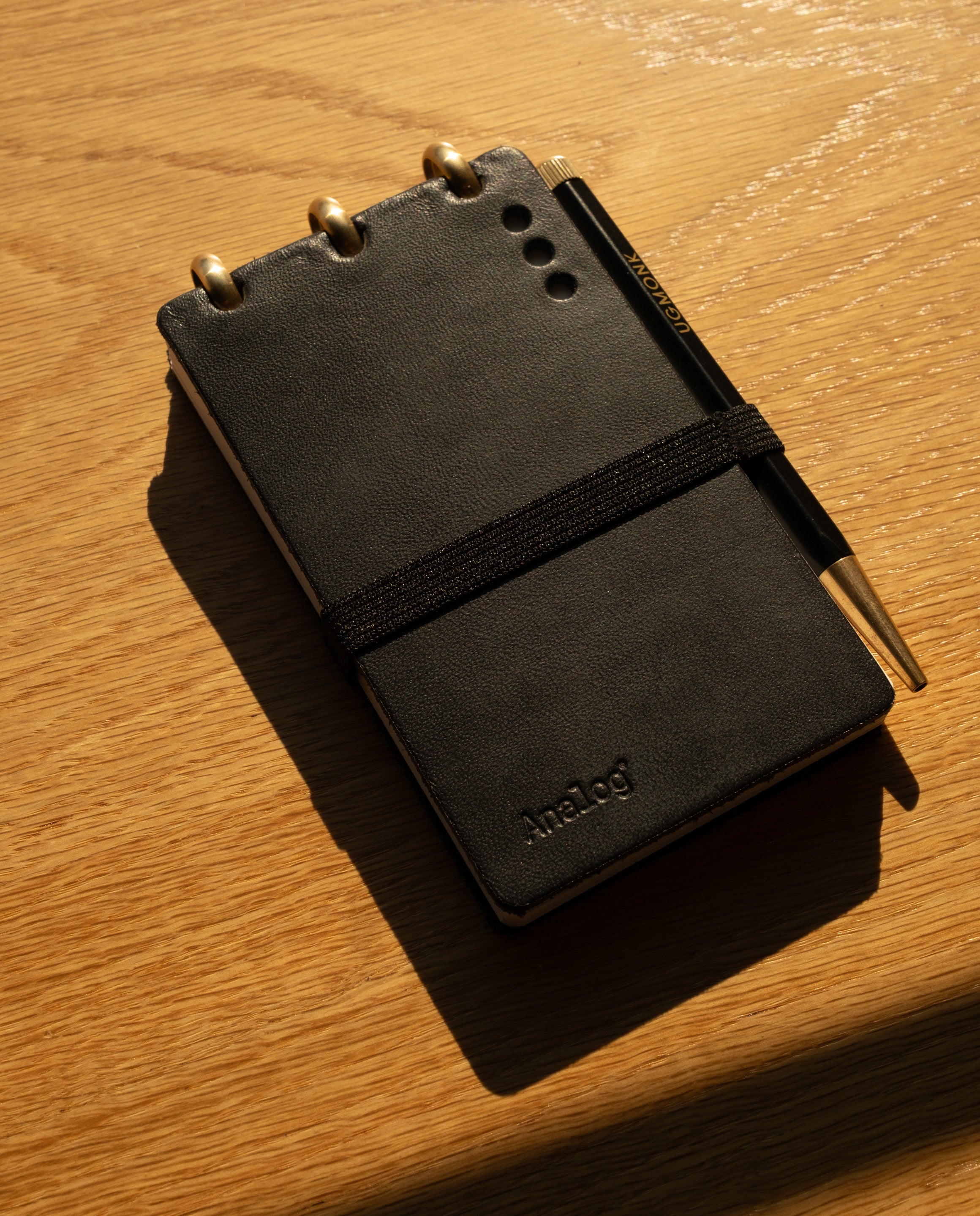 A Leather Discbound Analog + 3 Refills (Black) notebook, featuring full-grain black leather covers and an elastic band, sits on a wooden surface in warm light. A matching black pen rests on top, with Analog Task Cards visible inside.