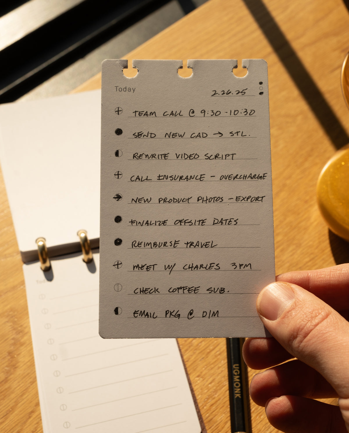 A hand holds a to-do list featuring tasks like team call, sending CAD files, rewriting the video script, calling insurance, reviewing product photos, finalizing dates, reimbursing travel expenses, meeting Charles, checking the coffee subscription, and emailing a package—all neatly organized on refills and protected by the elegant Leather Discbound Analog + 3 Refills (Tan) with its sophisticated full-grain leather cover.