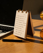 A compact Leather Discbound Analog (Brown) notebook with full-grain leather covers rests on a desk beside a pen and laptop, illuminated by warm sunlight. Inside, the to-do list features tasks such as calls, scriptwriting, photography, and checking emails—making it the ideal companion for organizing your day.