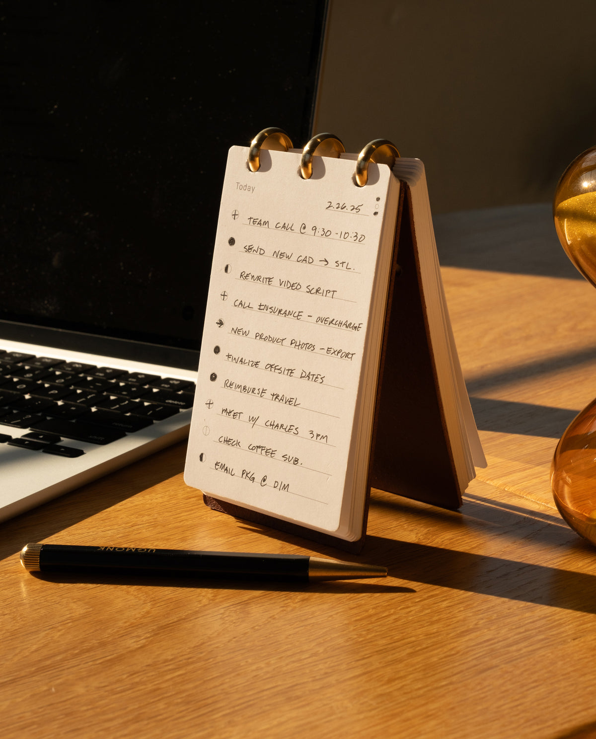 A compact Leather Discbound Analog (Brown) notebook with full-grain leather covers rests on a desk beside a pen and laptop, illuminated by warm sunlight. Inside, the to-do list features tasks such as calls, scriptwriting, photography, and checking emails—making it the ideal companion for organizing your day.