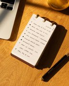 A Leather Discbound Analog (Brown) rests on a wooden desk, its full-grain leather covers safeguarding a handwritten to-do list, accompanied by a black pen, a laptop, and a glass of iced drink—all illuminated by warm sunlight.