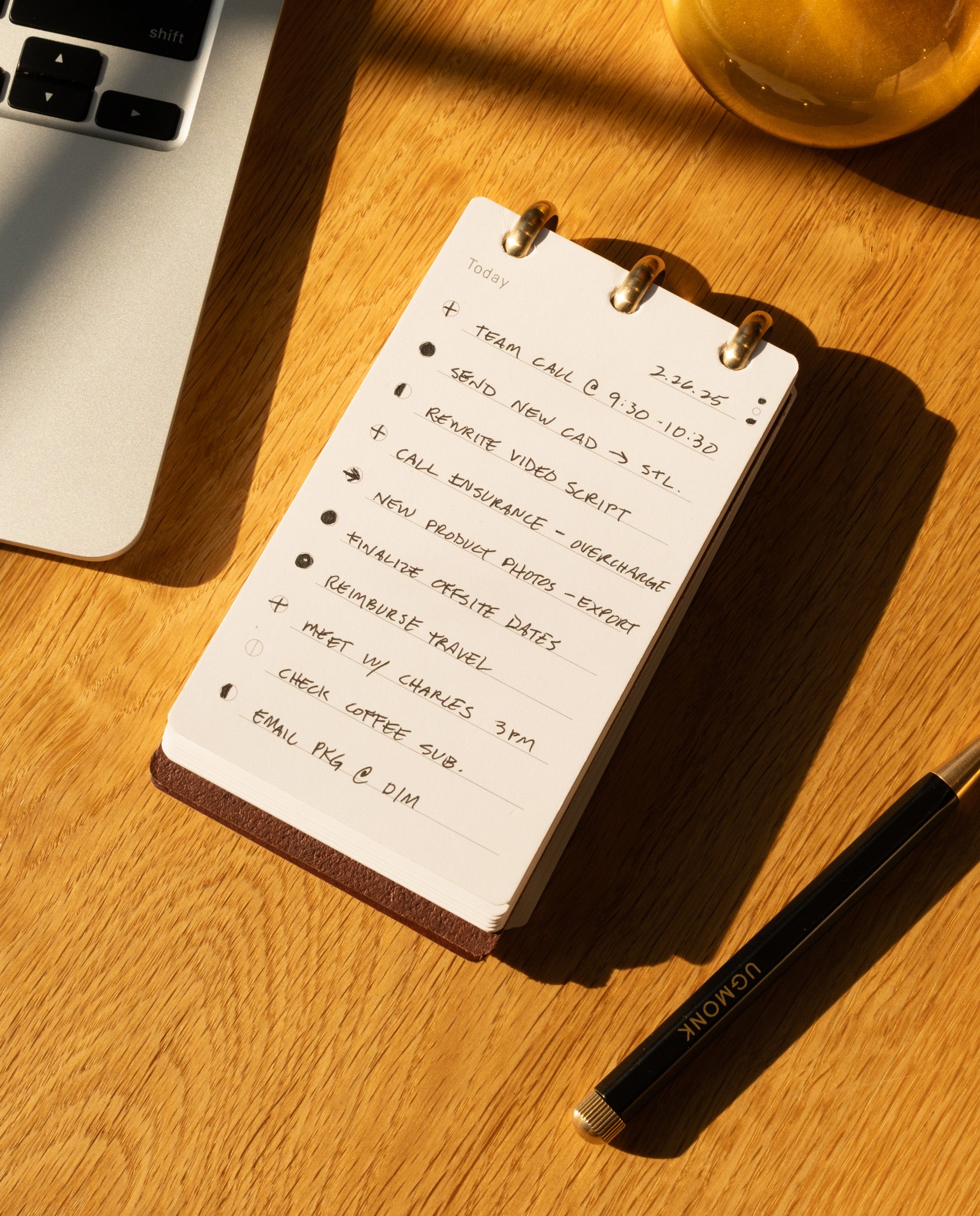 A Leather Discbound Analog (Brown) rests on a wooden desk, its full-grain leather covers safeguarding a handwritten to-do list, accompanied by a black pen, a laptop, and a glass of iced drink—all illuminated by warm sunlight.