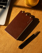 A Leather Discbound Analog (Brown), a black and gold pen, and part of a laptop rest on a wooden desk bathed in warm sunlight—capturing the refined appeal of Discbound Analog systems.