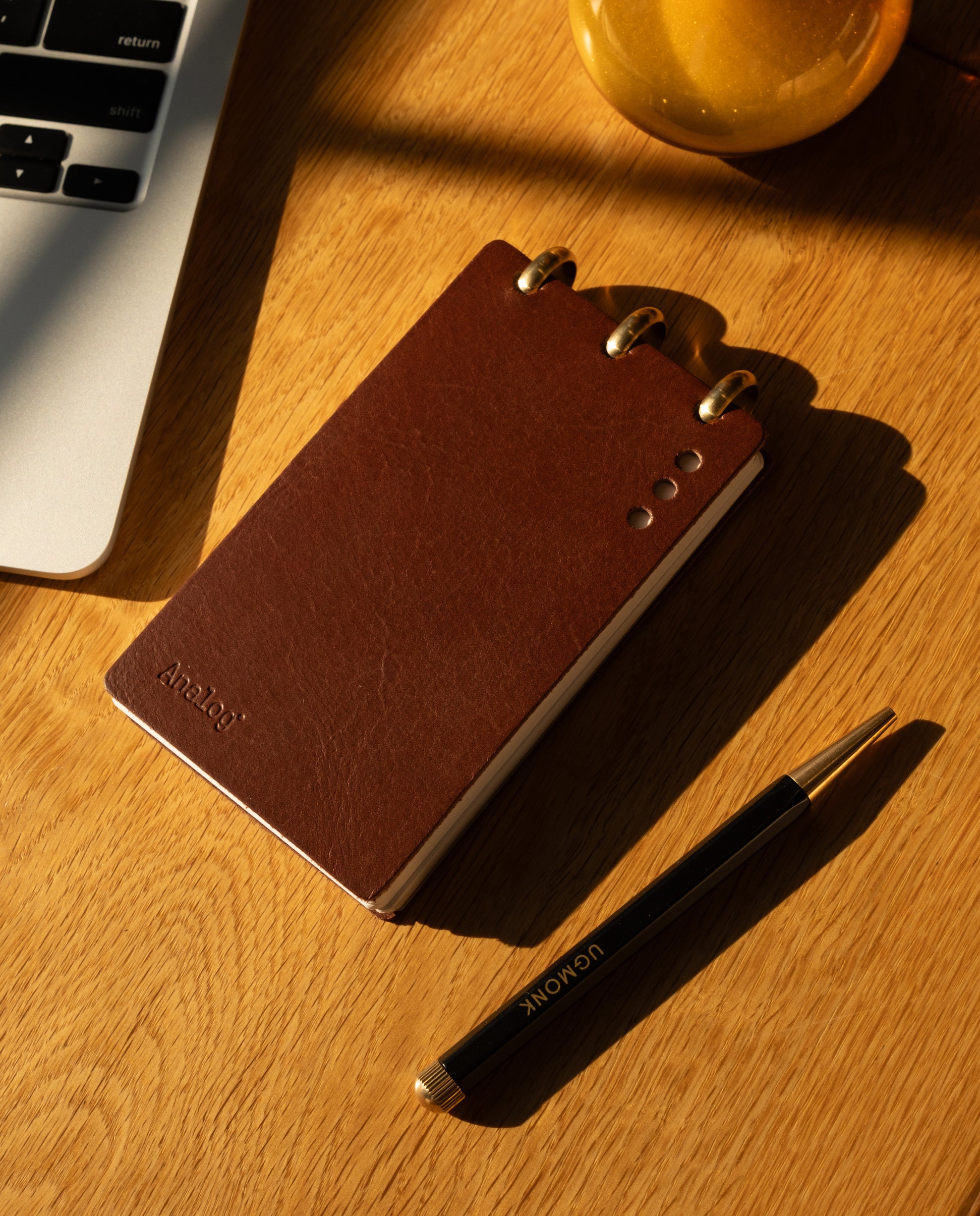 A Leather Discbound Analog (Brown), a black and gold pen, and part of a laptop rest on a wooden desk bathed in warm sunlight—capturing the refined appeal of Discbound Analog systems.