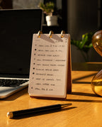 A Leather Discbound Analog (Brown) notepad with a handwritten to-do list sits on a wooden desk next to a black pen, bathed in warm sunlight. The list, organized on Analog Task Cards, includes tasks like team call, rewrite video script, and email FAQ.