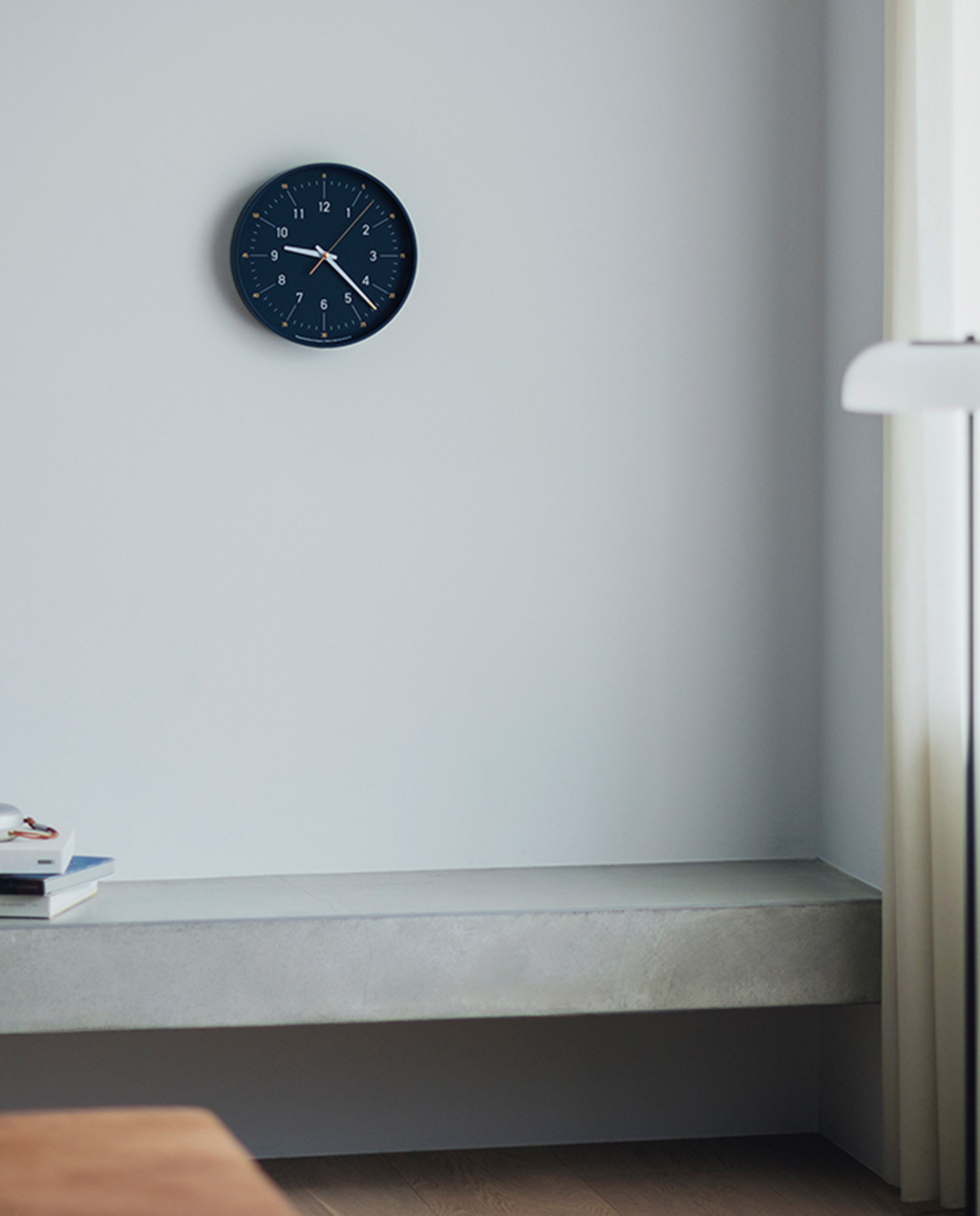 A Lemnos Essent Wall Clock (Navy) showing 11:20 hangs above a minimalist bench in a bright room, with a stack of books and sunlight streaming through the window.