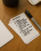 A stack of Analog Lined Cards - Small sits on a wooden table, accompanied by a Sharpie marker. The top card, one of the neatly ruled cards from the stack, lists groceries: eggs, sourdough, sharp cheddar, tomato sauce, cream cheese, red onion chips, tortillas, soup, clementines, and cake mix.