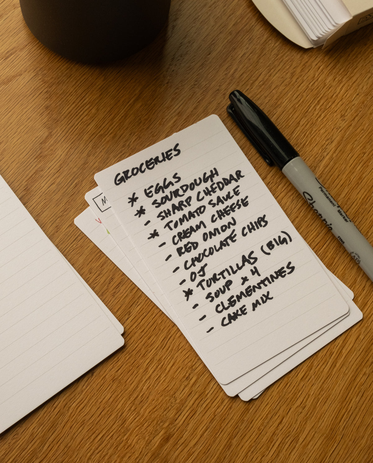 A stack of Analog Lined Cards - Small sits on a wooden table, accompanied by a Sharpie marker. The top card, one of the neatly ruled cards from the stack, lists groceries: eggs, sourdough, sharp cheddar, tomato sauce, cream cheese, red onion chips, tortillas, soup, clementines, and cake mix.