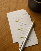 A stack of Analog Lined Cards - Small rests on a wooden table, with the top card featuring handwritten notes outlining a web roadmap, its stages highlighted in green and orange. A silver pen lies atop the ruled index cards next to a dark round object.
