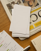 A stack of Analog Lined Cards - Small sits on a wooden table beside a folded newspaper, a notebook with handwritten notes, a silver pen, and a beige Analog-branded box.