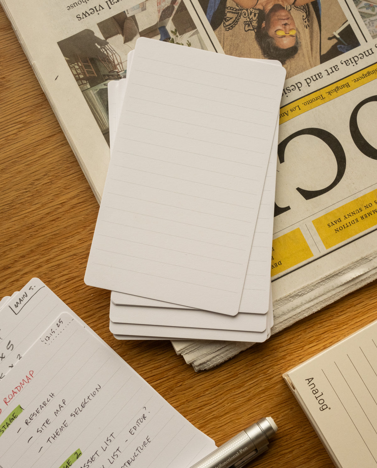 A stack of Analog Lined Cards - Small sits on a wooden table beside a folded newspaper, a notebook with handwritten notes, a silver pen, and a beige Analog-branded box.