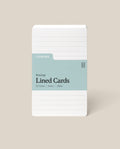 A stack of Analog Lined Cards - Small, in white 3x5 with a blue-green label, 50 cards made from 100lb uncoated cardstock, displayed upright on a beige background.