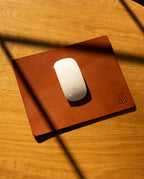A white wireless mouse sits on the Ugmonk Leather Mousepad (Tan) atop a wooden surface, diagonal shadows falling across the full-grain leather.