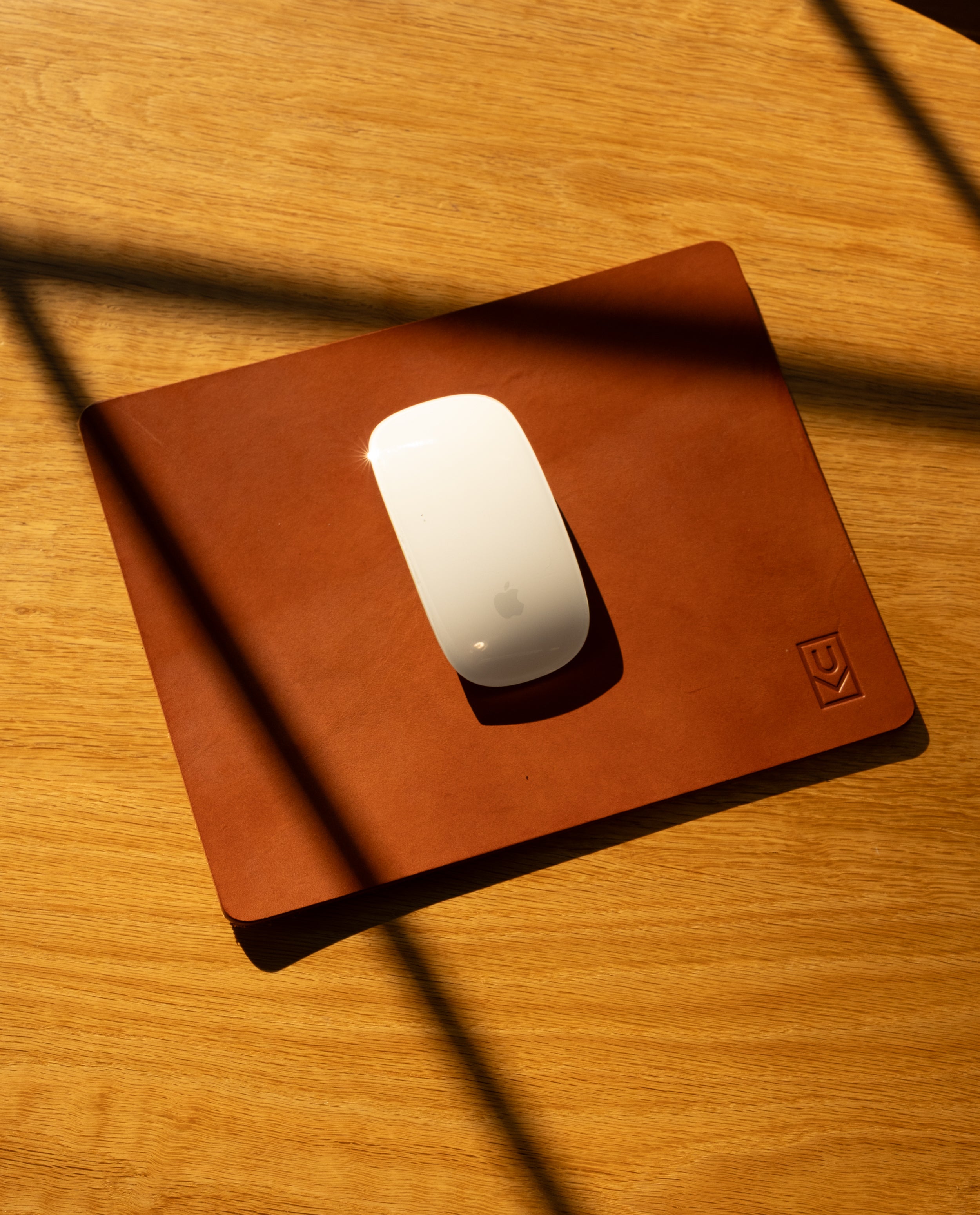 A white wireless mouse sits on the Ugmonk Leather Mousepad (Tan) atop a wooden surface, diagonal shadows falling across the full-grain leather.