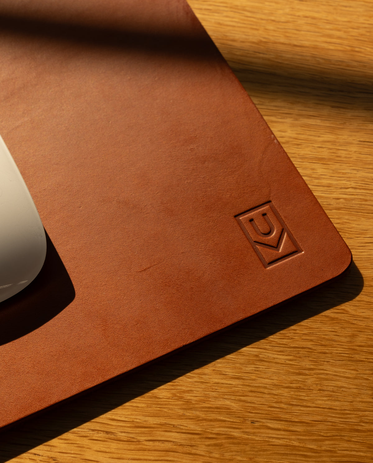 A close-up of the Ugmonk Leather Mousepad (Tan), crafted from brown leather with a geometric logo embossed in the corner, rests on wood. A white computer mouse is partly visible as sunlight casts gentle shadows across the scene.