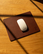 A white Apple Magic Mouse rests on the Ugmonk Leather Mousepad (Brown), handcrafted and set on a wooden desk where diagonal shadows fall across the surface.