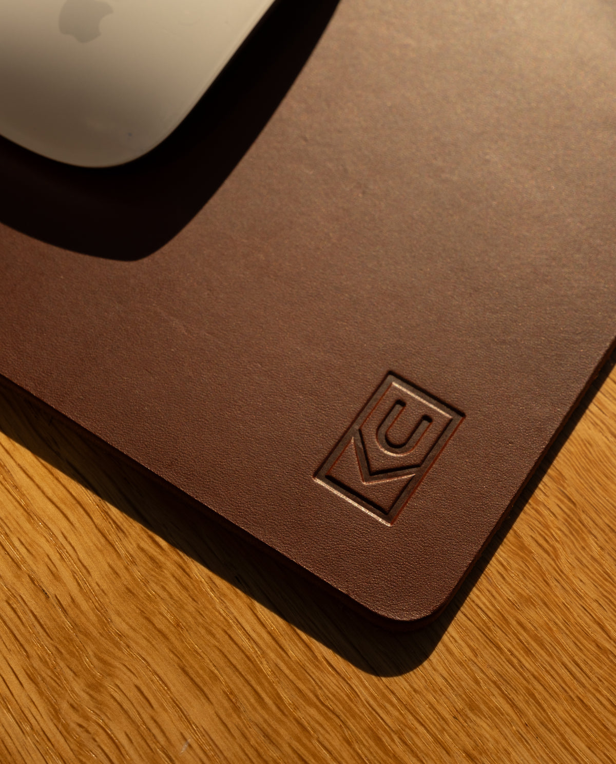 A close-up of the Ugmonk Leather Mousepad (Brown) with an embossed logo in the corner, placed on a wooden surface. The edge of a white computer mouse appears at the top left.