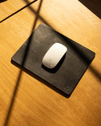 A white Apple Magic Mouse rests on an Ugmonk Leather Mousepad (Black), positioned on a light wooden desk, with sunlight casting diagonal shadows across the surface.