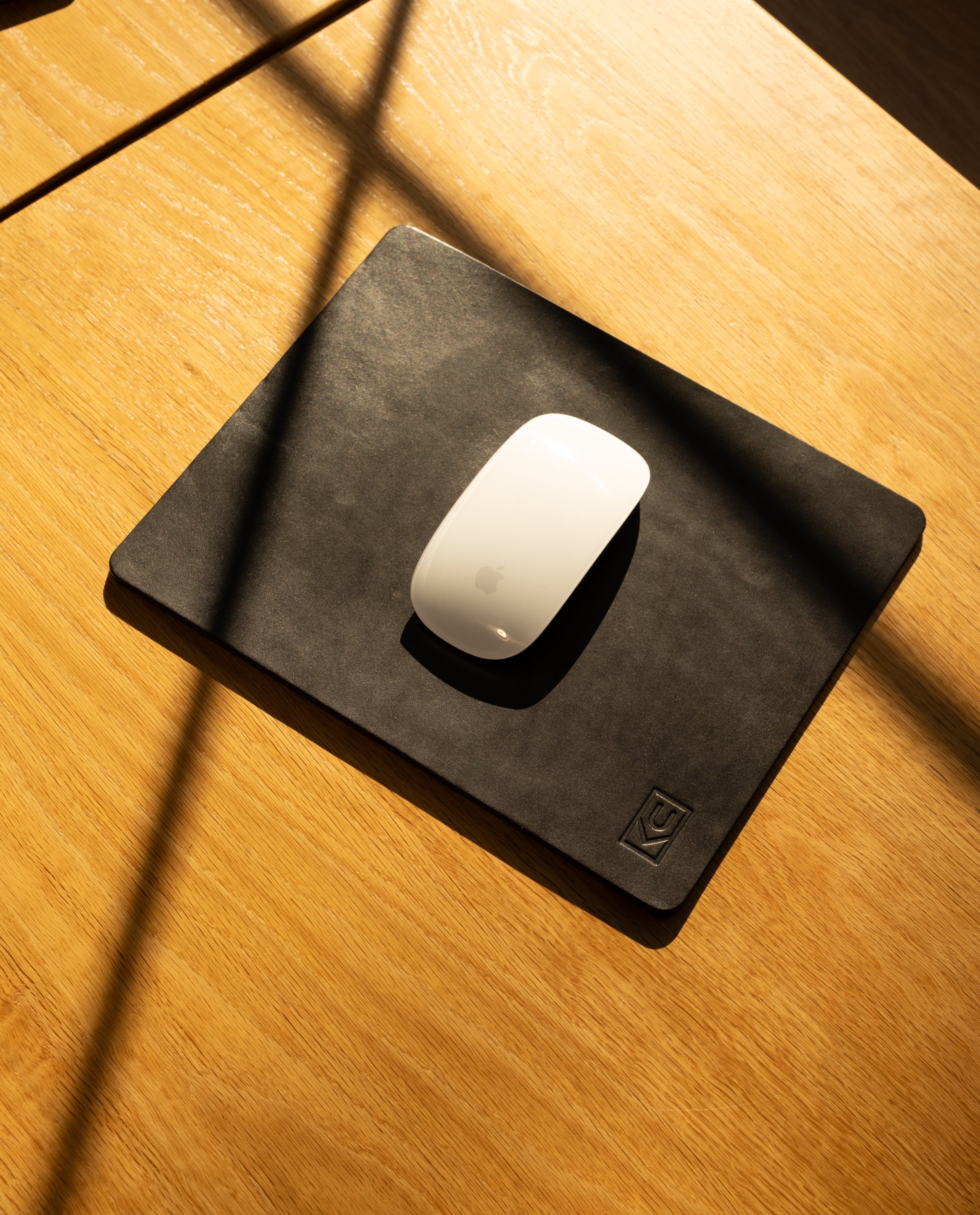 A white Apple Magic Mouse rests on an Ugmonk Leather Mousepad (Black), positioned on a light wooden desk, with sunlight casting diagonal shadows across the surface.