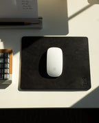 A white Apple Magic Mouse rests on an Ugmonk Leather Mousepad (Black) atop a white desk, next to a partially visible keyboard and a notepad, with sunlight casting shadows across the surface.