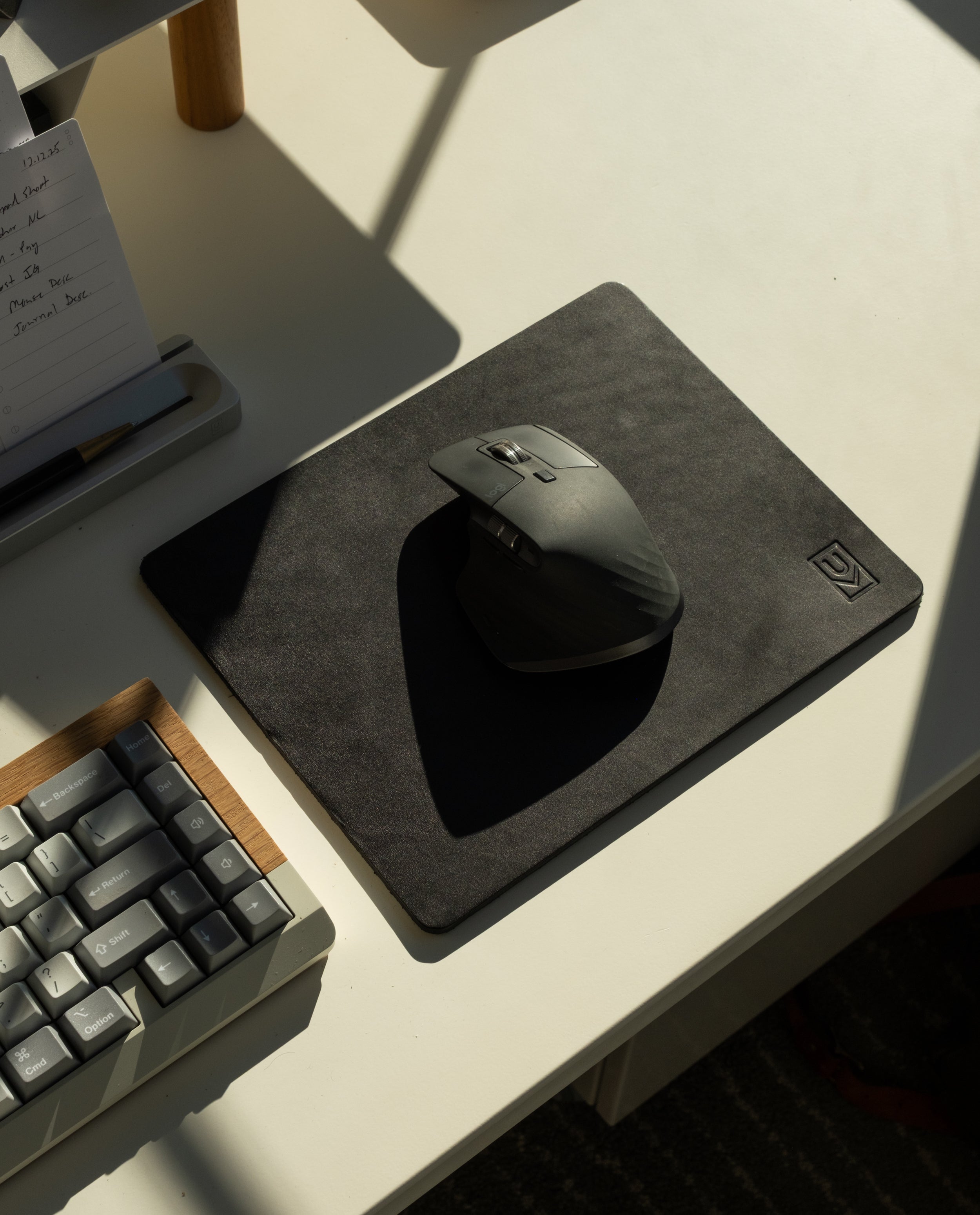A black computer mouse rests on the Ugmonk Leather Mousepad (Black), placed beside a gray keyboard and a notepad on a white desk, all illuminated by natural sunlight.