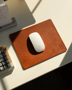 A white wireless mouse sits on the Ugmonk Leather Mousepad (Tan) on a white desk, next to a gray keyboard and notepad. Sunlight casts gentle shadows across the workspace.