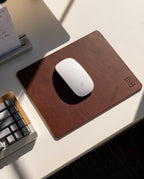 A white Apple Magic Mouse sits on a brown Ugmonk Leather Mousepad, next to a gray keyboard and notepad on a sunlit white desk.