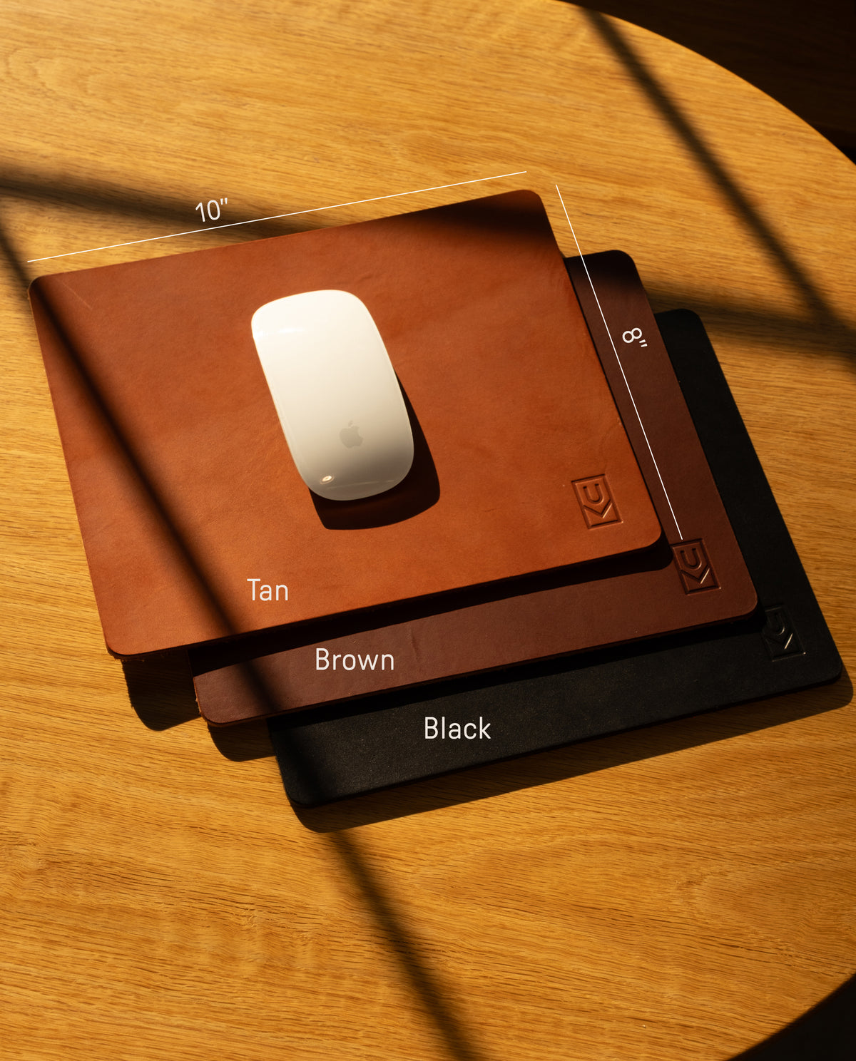 The Ugmonk Leather Mousepad (Tan), crafted from full-grain leather and sized 10" x 8", is shown on a wooden surface beside a white Apple Magic Mouse, catching sunlight and casting elegant shadows.
