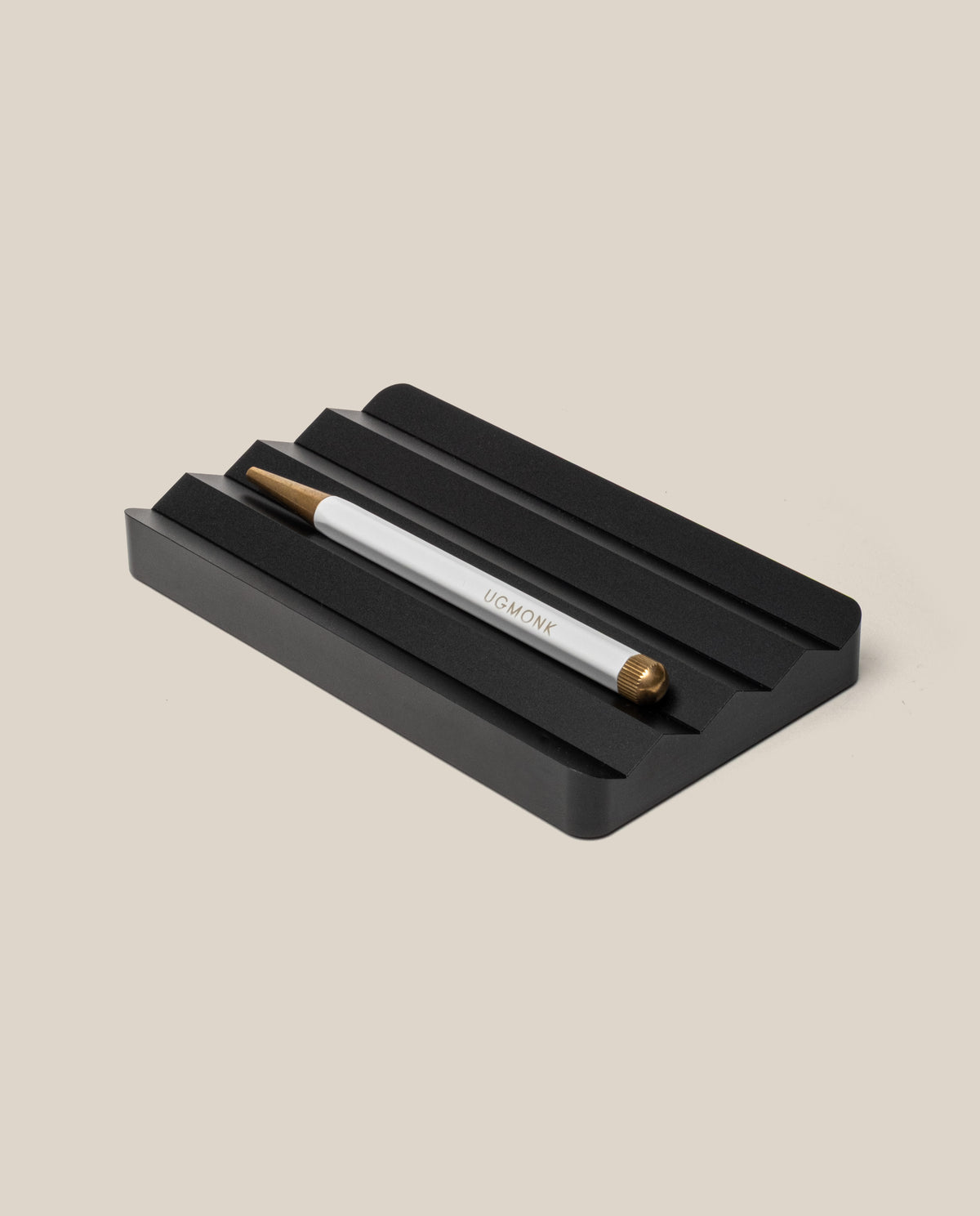 A silver and gold pen with "UGMONK" engraved rests in the Analog Multi Pen Tray (Black Aluminum) on a light beige background.