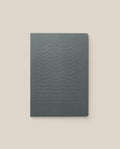 The Layflat Notebook (Charcoal) features a dark gray cover with embossed wavy lines, a minimalist design, dot grid pages, and Smyth sewn binding. It is shown on a light beige background.