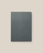 The Layflat Notebook (Charcoal) features a dark gray cover with an embossed wavy line pattern and Smyth sewn binding, shown against a light beige background.