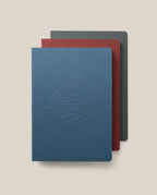 The Layflat Notebook Bundle (Series 2 - Set of 3) is stacked with blue on top (featuring a geometric embossed cover), red in the middle, and dark gray on the bottom; all have rounded corners.