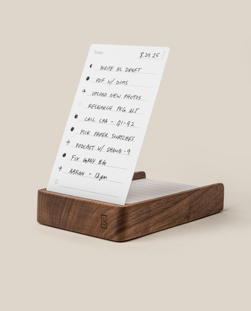 A wooden desk organizer holds a white notepad with a handwritten to-do list, including tasks like write ML draft, upload new photos, call CPA, and a meeting at noon with Aaron.