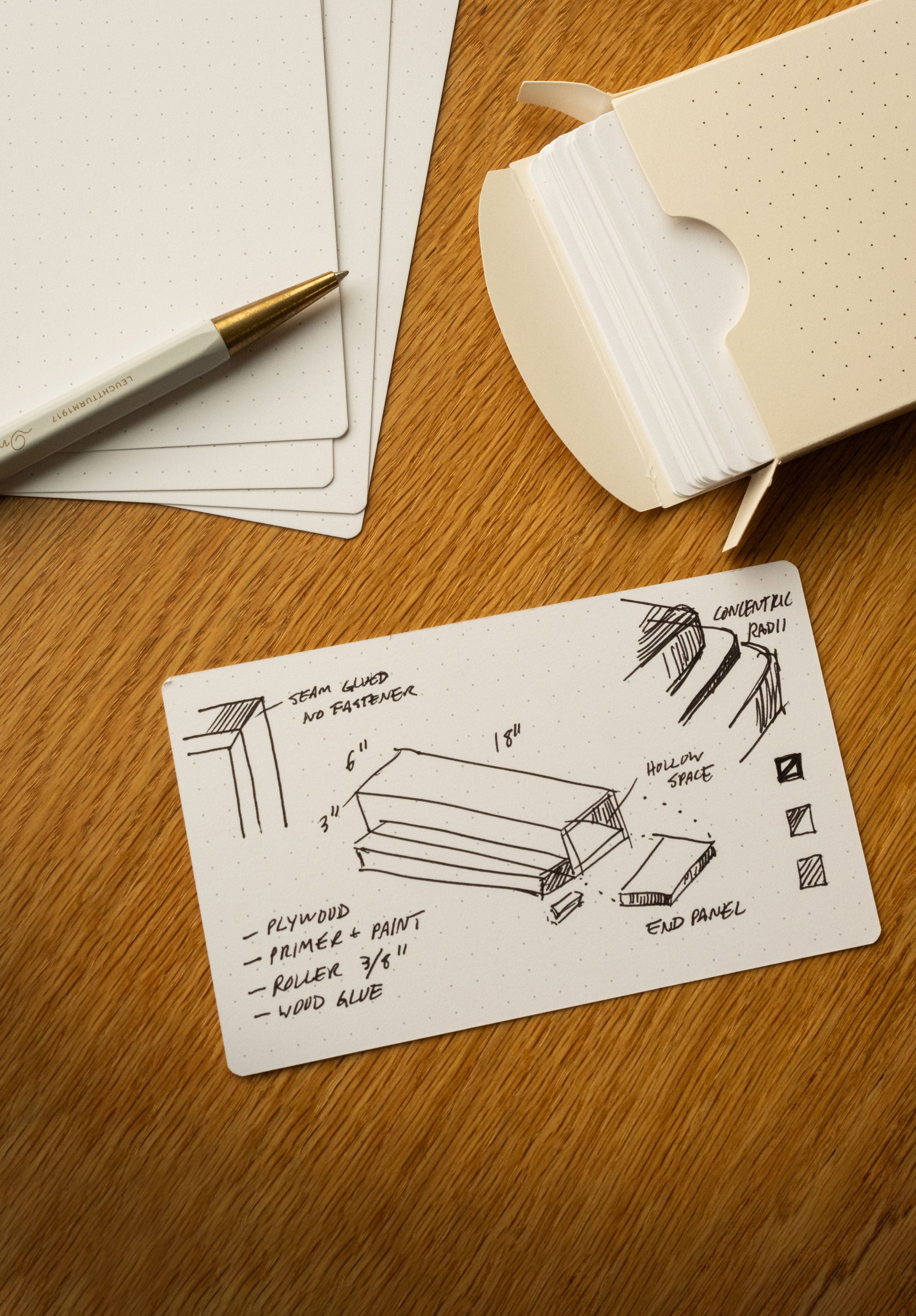 A pen, Analog Dot Grid Cards – Small, and a box are on a wooden desk. One 3x5 card made from sturdy 100lb card stock displays a sketch and notes for woodworking, including dimensions, materials, and construction details.
