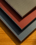 The Layflat Notebook Bundle (Series 2 - Set of 3) features gray, red, and teal dot grid notebooks with exposed Smyth sewn binding, resting on a wooden surface where sunlight highlights their textured paper and covers.