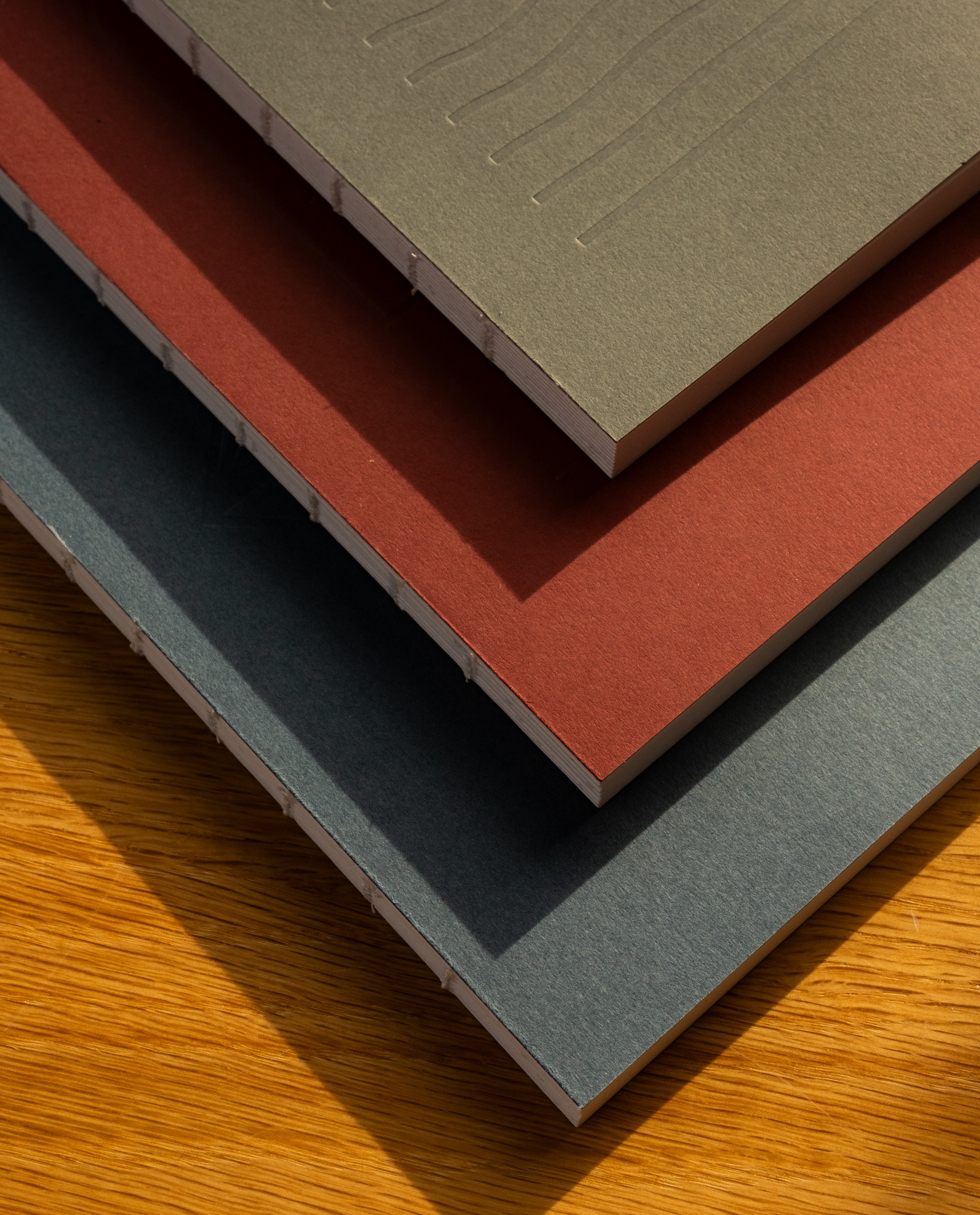 The Layflat Notebook Bundle (Series 2 - Set of 3) features gray, red, and teal dot grid notebooks with exposed Smyth sewn binding, resting on a wooden surface where sunlight highlights their textured paper and covers.
