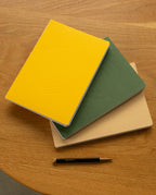 A closed Layflat Notebook (Yellow) with an embossed geometric cover design is stacked alongside green and beige notebooks on a wooden table. A black and gold pen rests beneath them. Each notebook is crafted to lay flat and features dot grid pages, making them ideal for effortless writing and sketching.