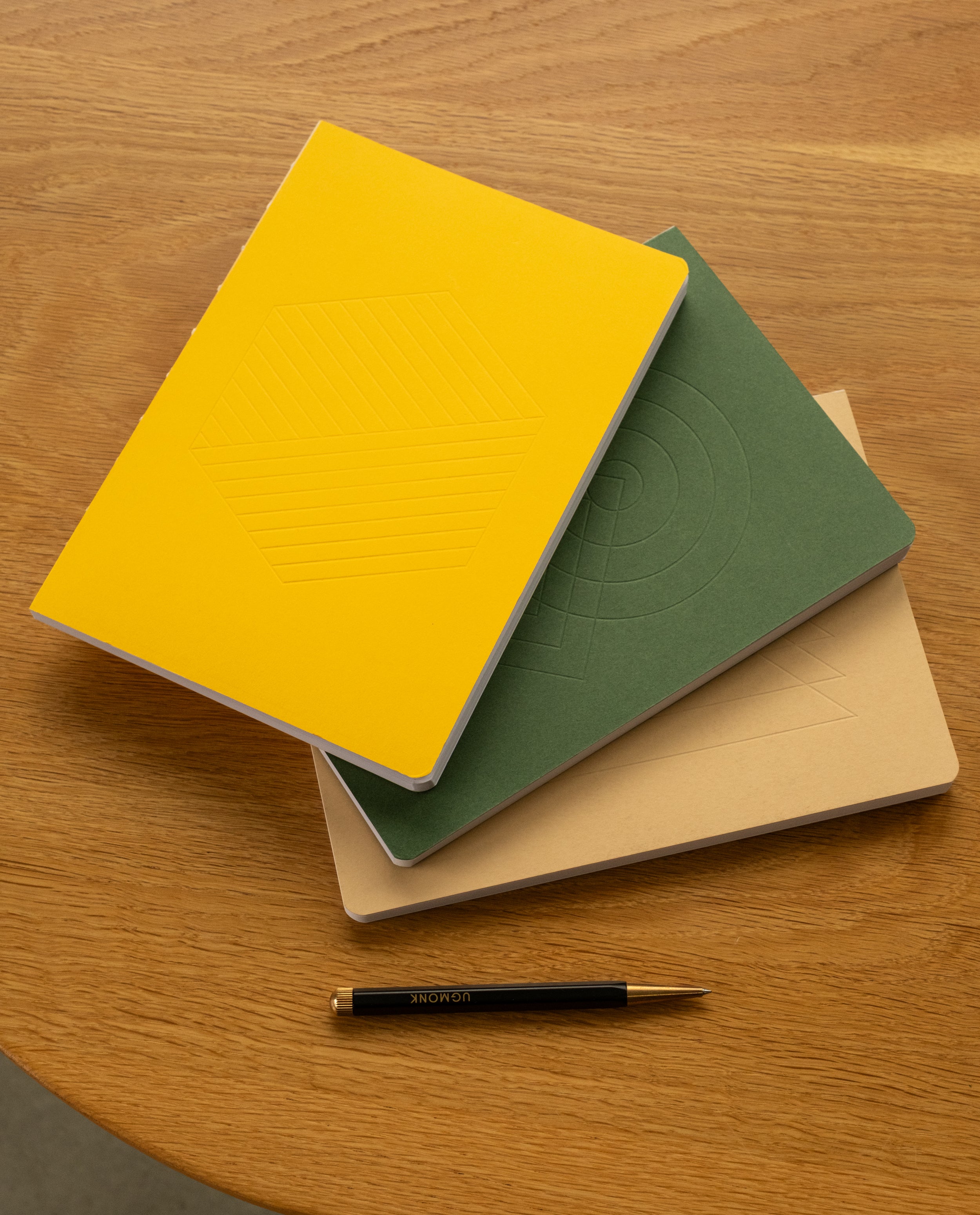 A closed Layflat Notebook (Yellow) with an embossed geometric cover design is stacked alongside green and beige notebooks on a wooden table. A black and gold pen rests beneath them. Each notebook is crafted to lay flat and features dot grid pages, making them ideal for effortless writing and sketching.