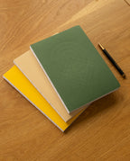 Three notebooks from the Layflat Notebook Bundle (Set of 3)—in green, beige, and yellow—are stacked on a wooden table next to a black and gold pen. The green notebook showcases an embossed geometric pattern on its cover, and each notebook in the set is designed with durable Smyth sewn binding for easy layflat writing.