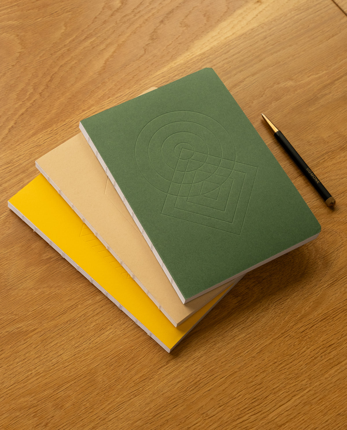 Three notebooks from the Layflat Notebook Bundle (Set of 3)—in green, beige, and yellow—are stacked on a wooden table next to a black and gold pen. The green notebook showcases an embossed geometric pattern on its cover, and each notebook in the set is designed with durable Smyth sewn binding for easy layflat writing.