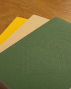 Three closed notebooks from the Layflat Notebook Bundle (Set of 3) are stacked on a wooden surface. The top notebook features a green geometric pattern, the middle is beige, and the bottom is yellow.