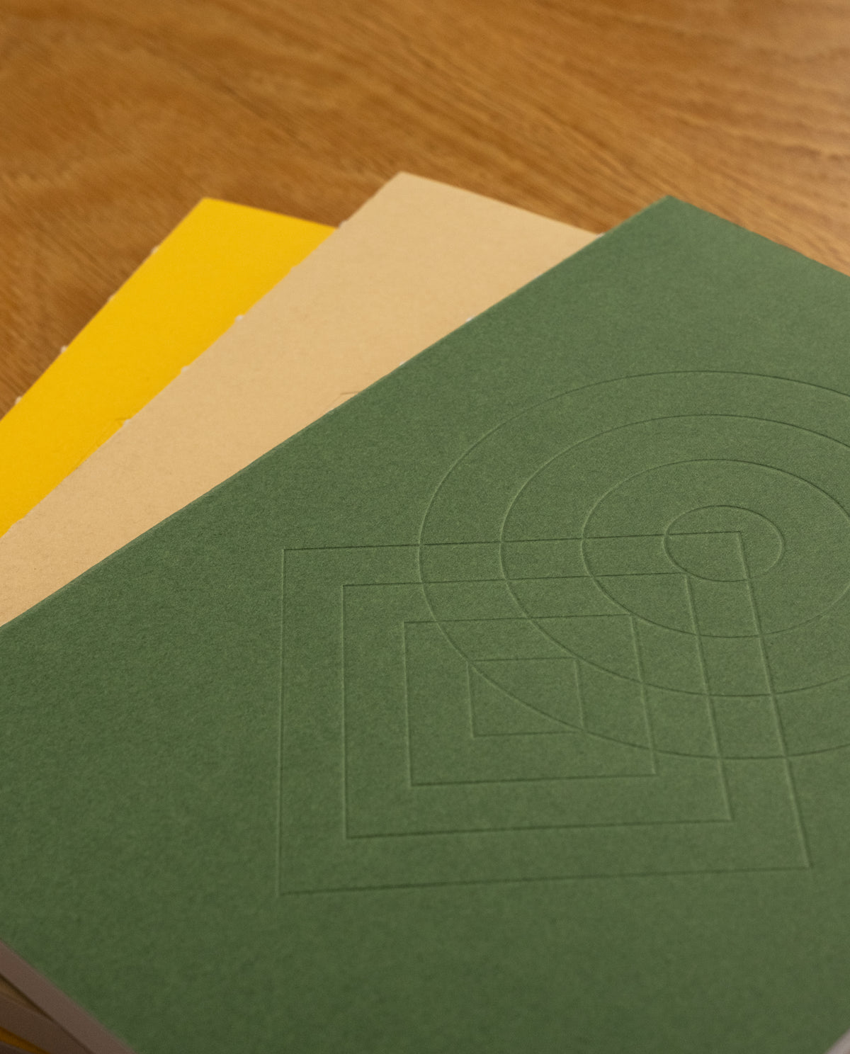 Three closed notebooks from the Layflat Notebook Bundle (Set of 3) are stacked on a wooden surface. The top notebook features a green geometric pattern, the middle is beige, and the bottom is yellow.
