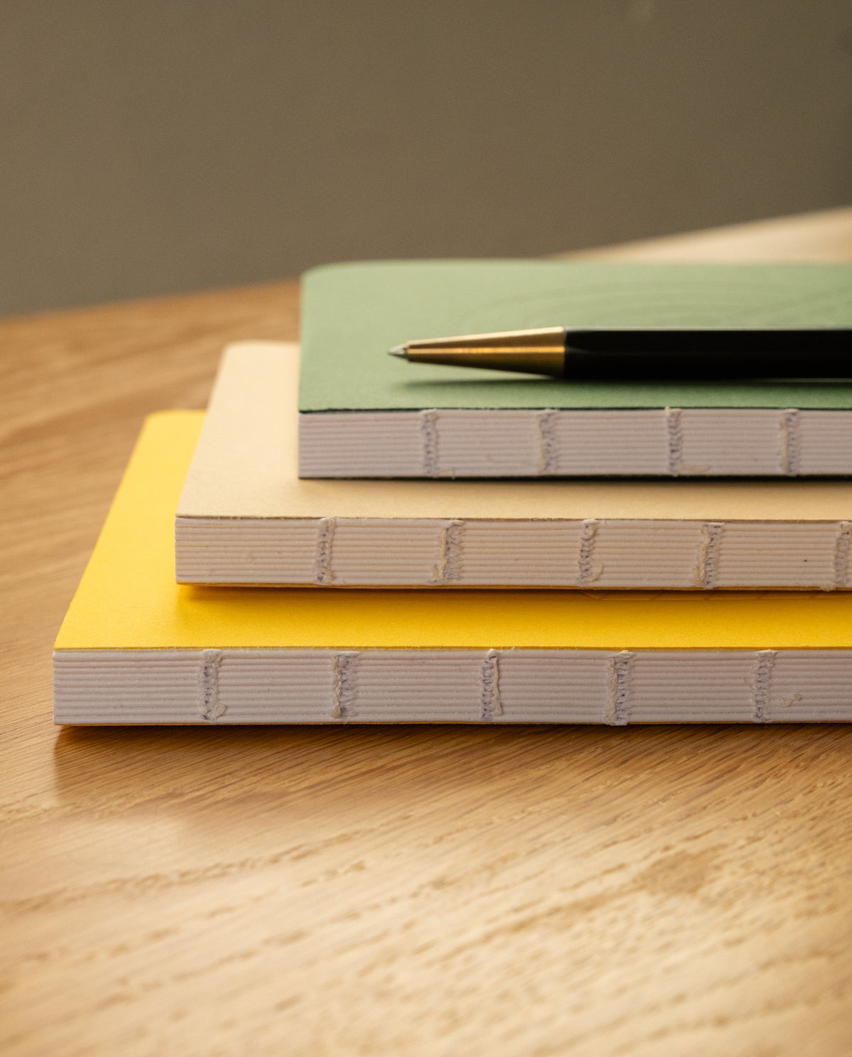 Three Layflat Notebooks, stacked on a wooden surface, showcase green, beige, and yellow covers. Each features Smyth sewn binding for enhanced durability and convenient lay-flat use, with dot grid pages perfect for organized notes or sketches. A black and gold pen rests atop the yellow Layflat Notebook.