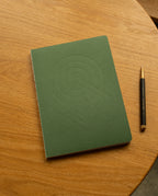 A Layflat Notebook (Green) with geometric embossed shapes on the cover lies on a round wooden table next to a black and gold pen, its Smyth sewn binding ensuring it opens flat for effortless writing.
