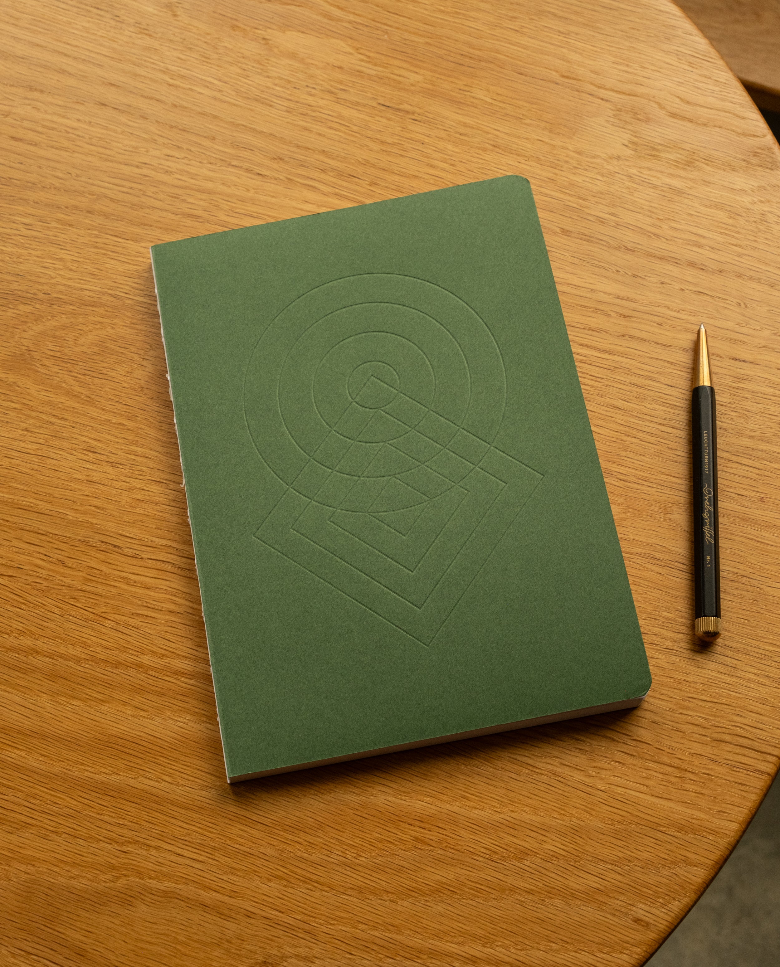 A Layflat Notebook (Green) with geometric embossed shapes on the cover lies on a round wooden table next to a black and gold pen, its Smyth sewn binding ensuring it opens flat for effortless writing.