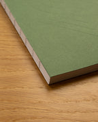 A close-up of the Layflat Notebook (Green) featuring an exposed spine binding and subtle embossed lines on its plain cover, resting on a light wooden surface.