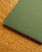 A Layflat Notebook (Green) with UGMONK embossed on the cover and Smyth sewn binding rests on a wooden surface.